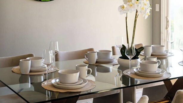 Un comedor con una mesa de vidrio, platos blancos y un jarrón de flores blancas.