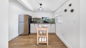 Una cocina compacta con un refrigerador de acero inoxidable, un microondas y una cafetera sobre la encimera. Hay un estante de madera con cuencos y tazas.