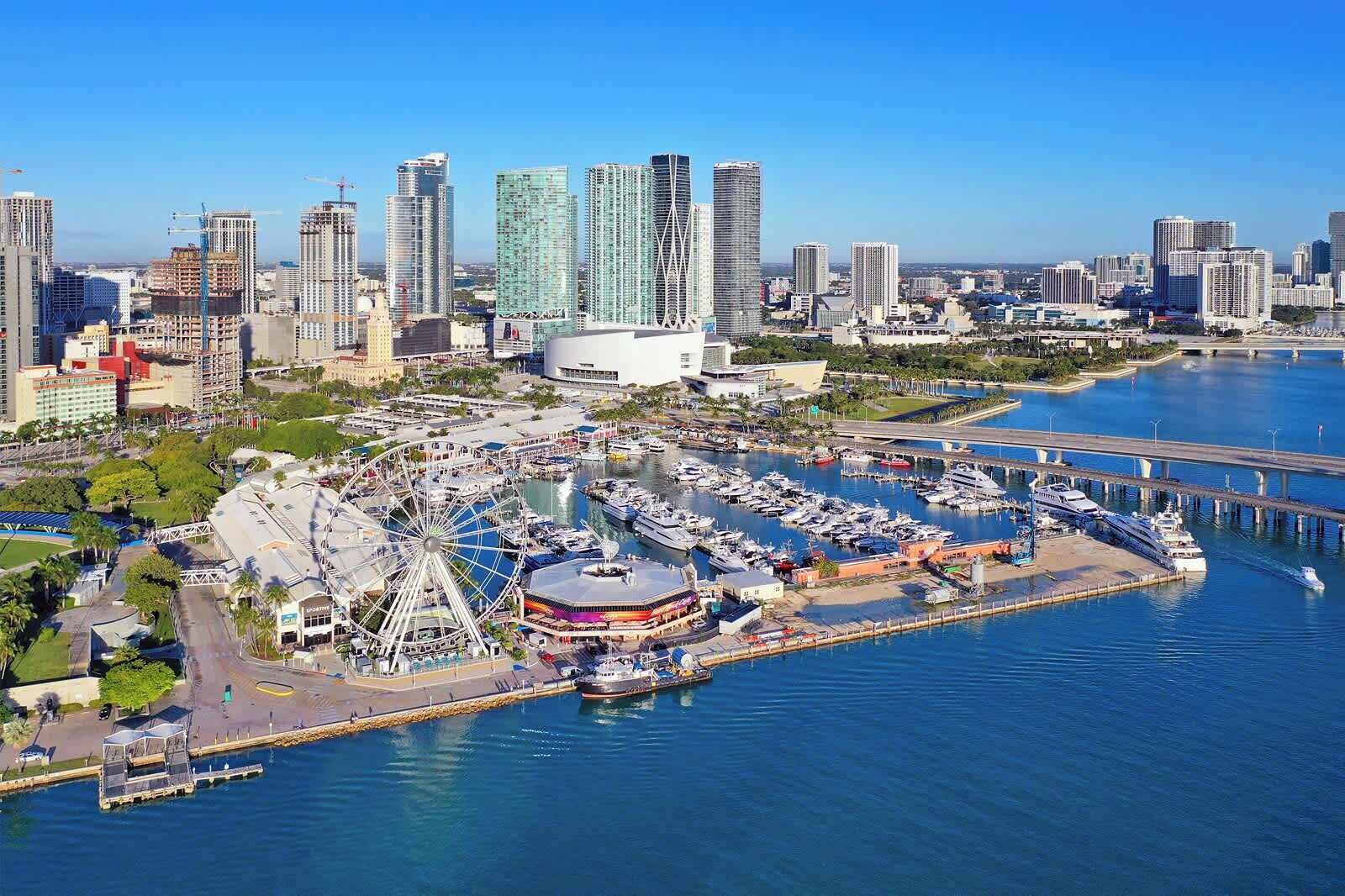 An aerial view of a marina with boats docked, a Ferris wheel, and modern high-rise buildings in the background.