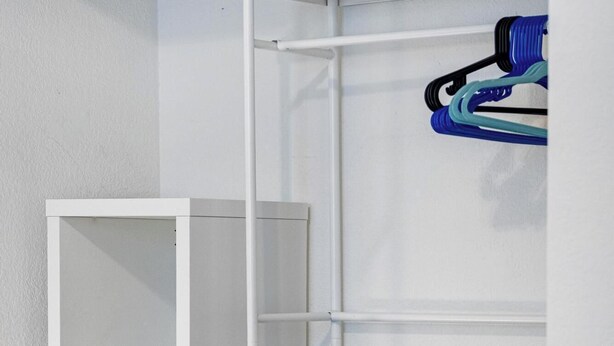 Een witte garderobe met planken en hangruimte, een klein strijkplank met een stoomstrijkijzer, en een blauw en zwart voorwerp aan de muur.