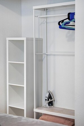 Un armario blanco con estantes y espacio para colgar, una plancha y tabla de planchar, y un objeto azul y negro colgado en la pared.