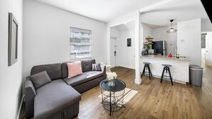 A modern living room with a grey sofa, a small round table with a vase, and a kitchen area with a bar counter and stools. - Art Deco Escape Steps to Ocean Free Parking (Miami Beach)
