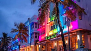 A brightly lit hotel with a neon sign, palm trees, and a street view. - Casa Mare 1BR Steps From the Ocean (Miami Beach)