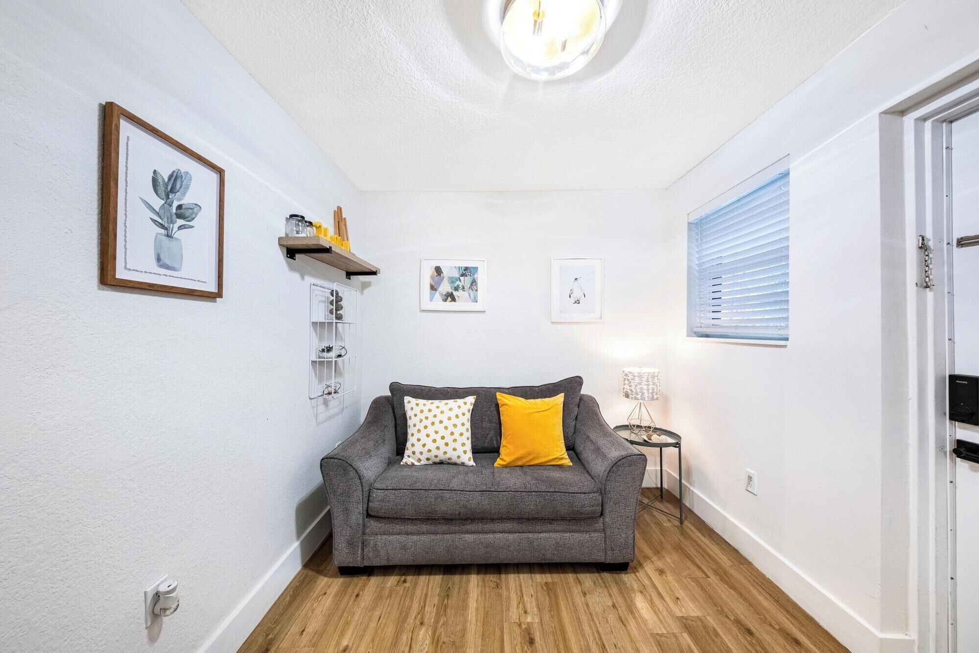 A cozy living room with a grey sofa, a small side table, and a lamp. There are framed pictures on the wall and a wooden shelf with decorative items.