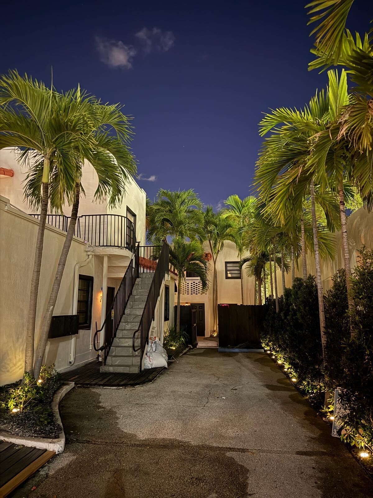A nighttime view of a courtyard with palm trees, a staircase, and a building facade.