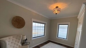 Una habitación con un sillón capitoné, una mesita de noche con una lámpara, un adorno en la pared, dos ventanas con vista a un cuerpo de agua y un candelabro.