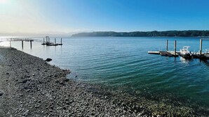 A pebbled beach with a wooden pier extending into calm waters, surrounded by clear skies and distant hills. - Hood Canal Waterfront - Dock Hot Tub BBQ Games (Union)