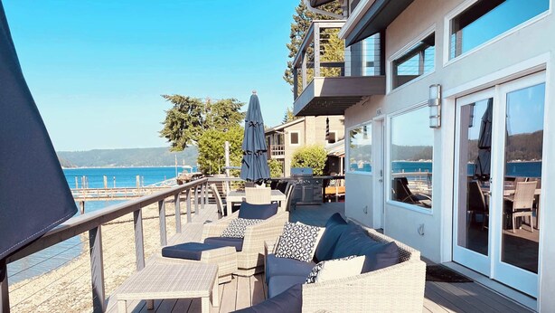 A deck with wicker furniture, a view of the sea, and a building with large windows. - Hood Canal Waterfront - Dock Hot Tub BBQ Games (Union)
