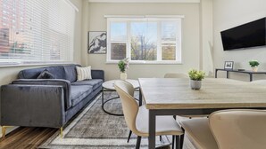 Ein modernes Wohnzimmer mit grauem Sofa, einem Esstisch mit Stühlen, einem an der Wand montierten Fernseher und einem Fenster mit Jalousien.