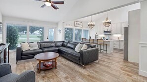 A spacious living room with a large L-shaped sectional sofa, a round coffee table, and a ceiling fan. The room features large windows with blinds, a kitchen area with white cabinets, and a chandelier. - Fore-Shore | Sawgrass CC Golf View Retreat | 3RB - 2BA | Walk to the Beach (Ponte Vedra Beach)
