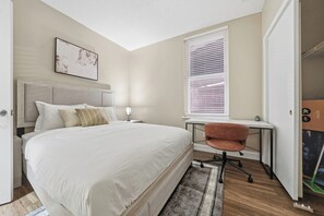 A bedroom with a bed, bedside table, lamp, chair, window, and a painting on the wall. - Home-style 1BR perfect for long visits (New Haven)
