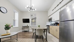 Une cuisine moderne avec un réfrigérateur en acier inoxydable, des armoires en bois, une table à manger avec des chaises, une horloge au mur et un téléviseur à écran plat fixé au-dessus du meuble télé.