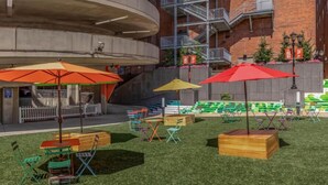 Un espace extérieur aménagé avec des chaises pliantes et des parasols colorés, entouré d’un bâtiment à la façade en briques et doté d’un escalier en colimaçon.