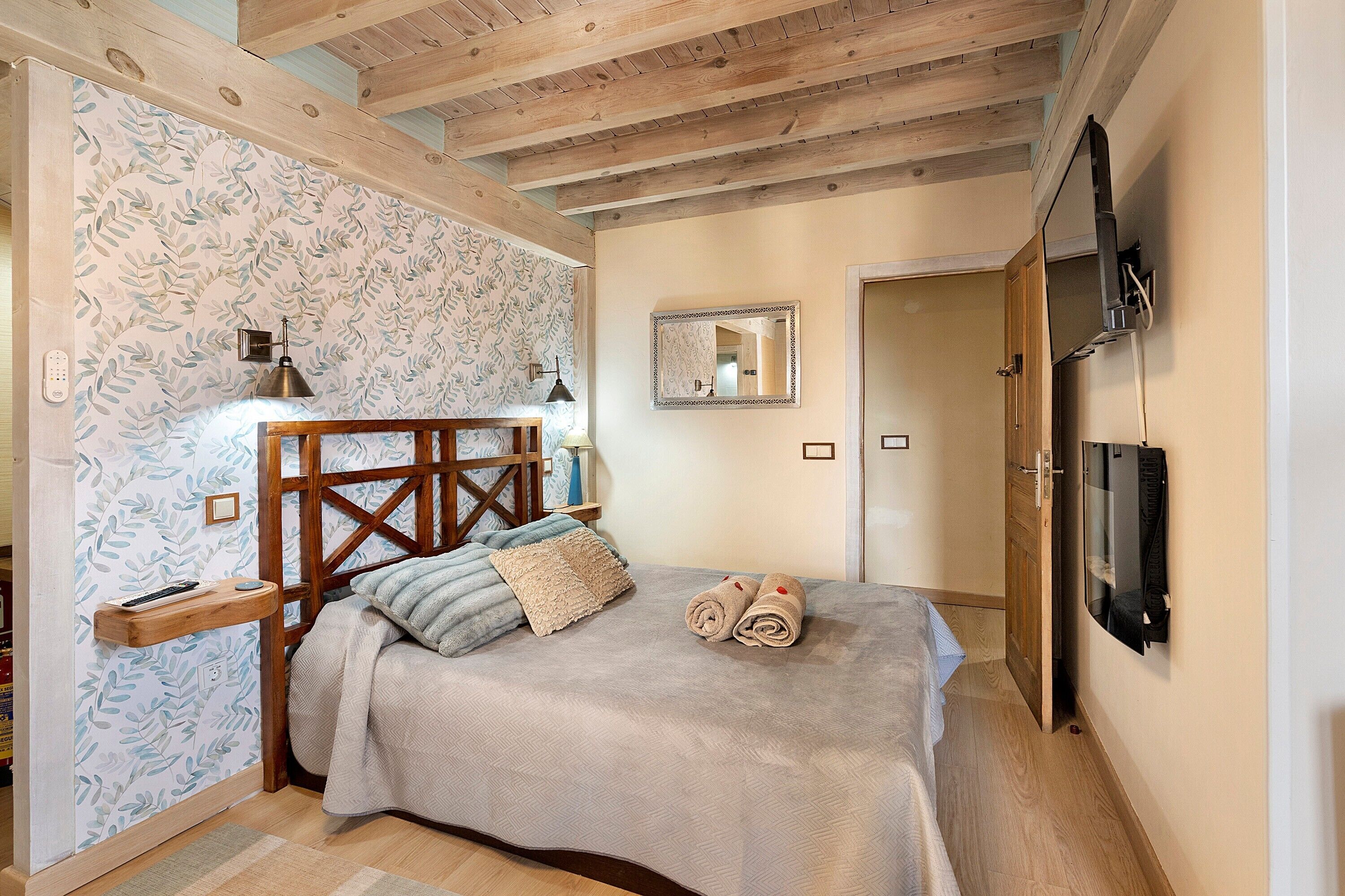 Een slaapkamer met een houten hoofdbord, een bed met een beige deken en kussens, een nachtkastje met een lamp en een aan de muur bevestigde televisie.