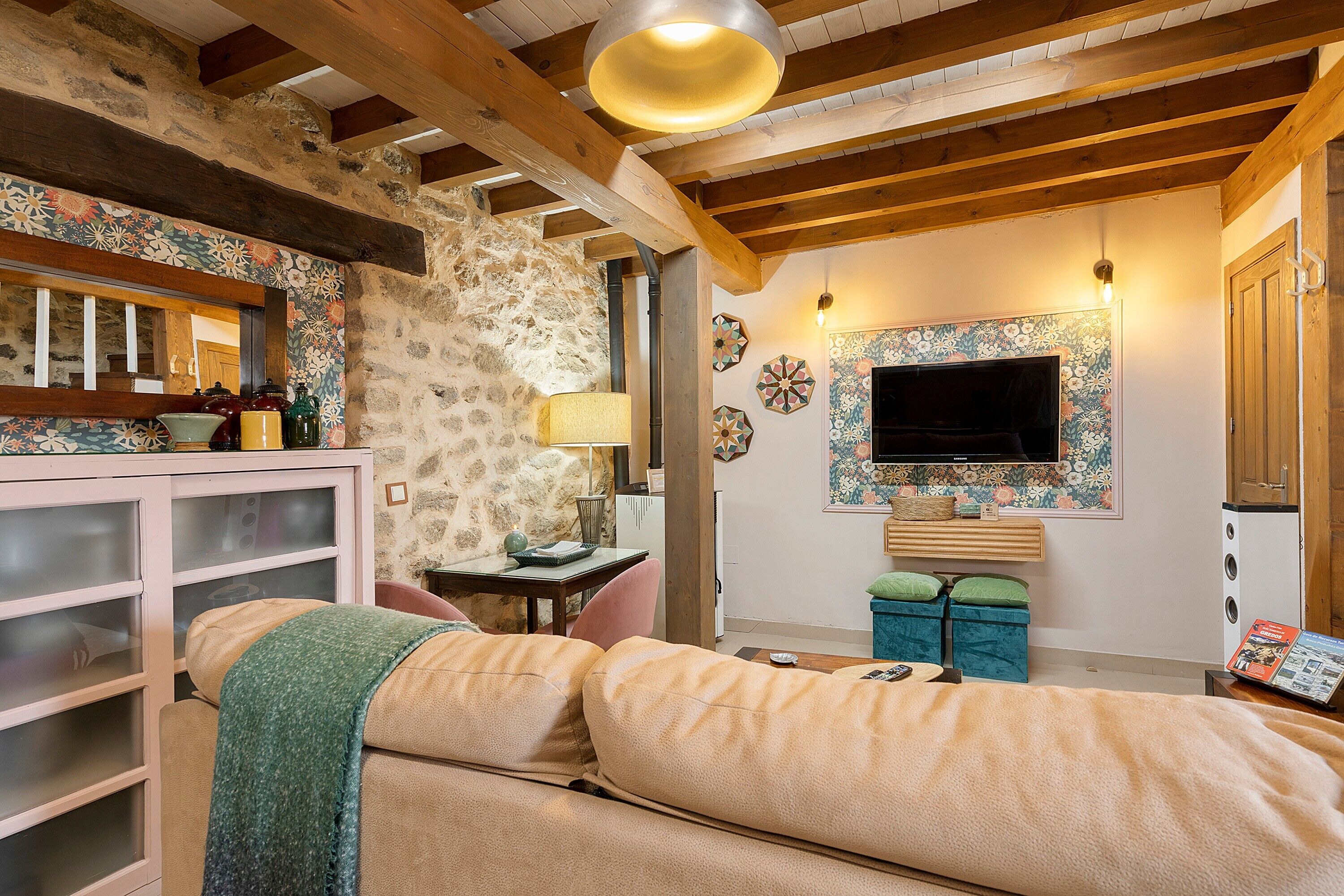 Uma sala de estar acolhedora com parede de pedra, vigas de madeira, um sofá, uma televisão e um armário com portas de vidro.