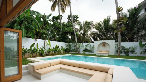 Un área de piscina moderna al aire libre con un sofá seccional color beige y una puerta de cristal que da a un patio.