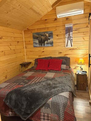 Un dormitorio acogedor tipo cabaña con una cama, un cuadro colgado de un alce, un calentador en la pared y una mesita con una lámpara.