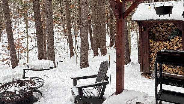 A covered outdoor seating area with wooden benches and a fire pit, surrounded by snow-covered trees.