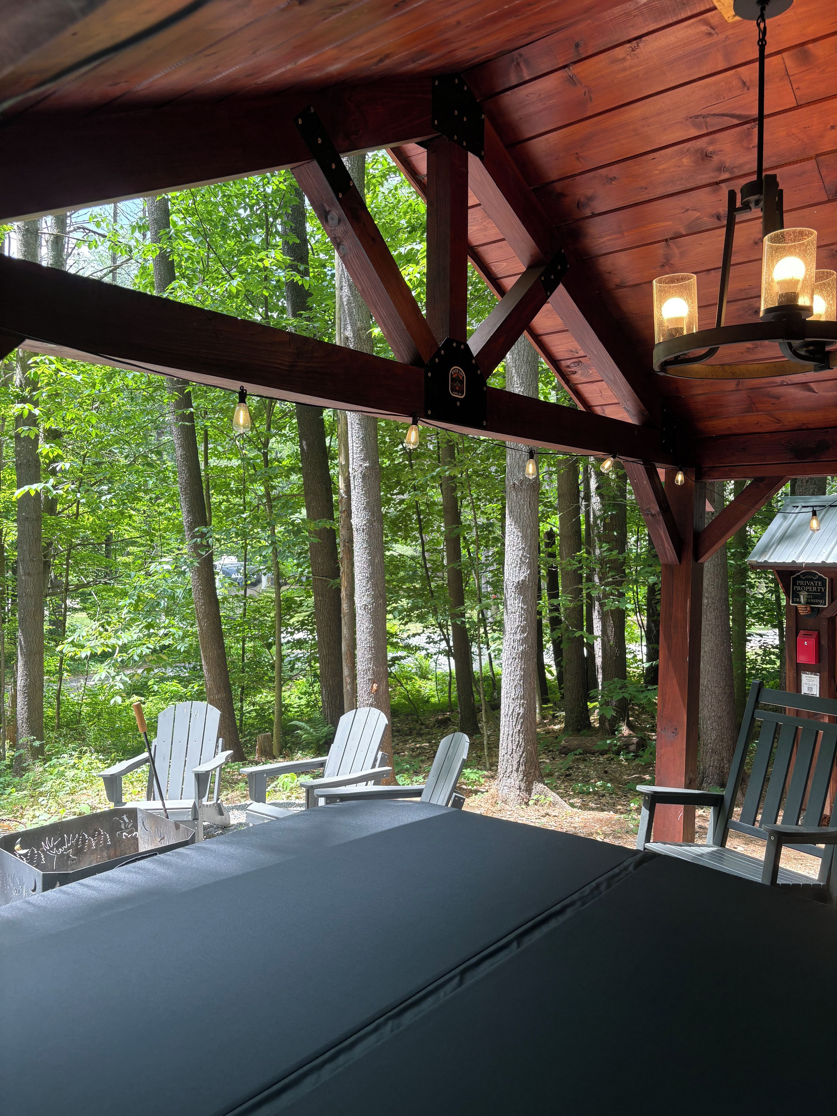 Een overdekt terras met Adirondack-stoelen en een tafel, omgeven door bomen.