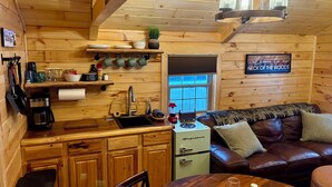 A cozy cabin interior with wooden walls, a kitchen area including a stove, sink, and coffee maker, and a living area with a couch and a dining table.