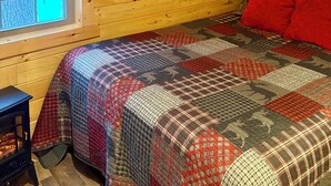 A cozy cabin room with wooden walls, a quilted bedspread, a window with blinds, and a painting of a moose.