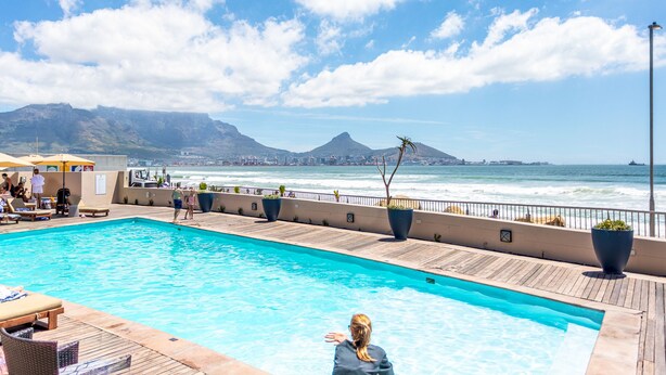 Una piscina con cubierta de madera y sillones de descanso, con vistas a una playa y al horizonte de la ciudad.