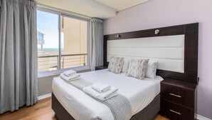 Una habitación de hotel con una cama grande, una mesita de noche de madera, una ventana con cortinas y vistas al mar.