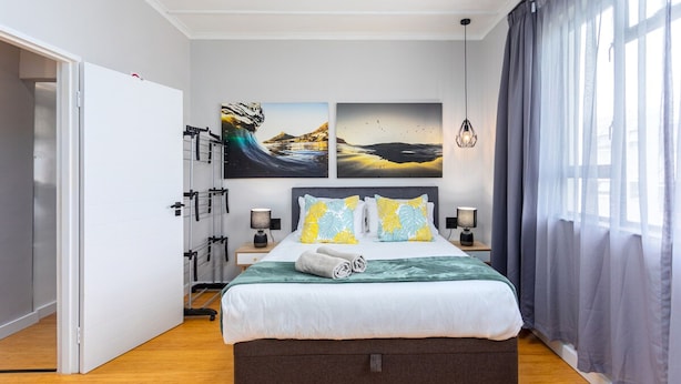 Quarto moderno com cama, duas luminárias de parede, uma janela grande com cortinas e duas imagens emolduradas na parede.