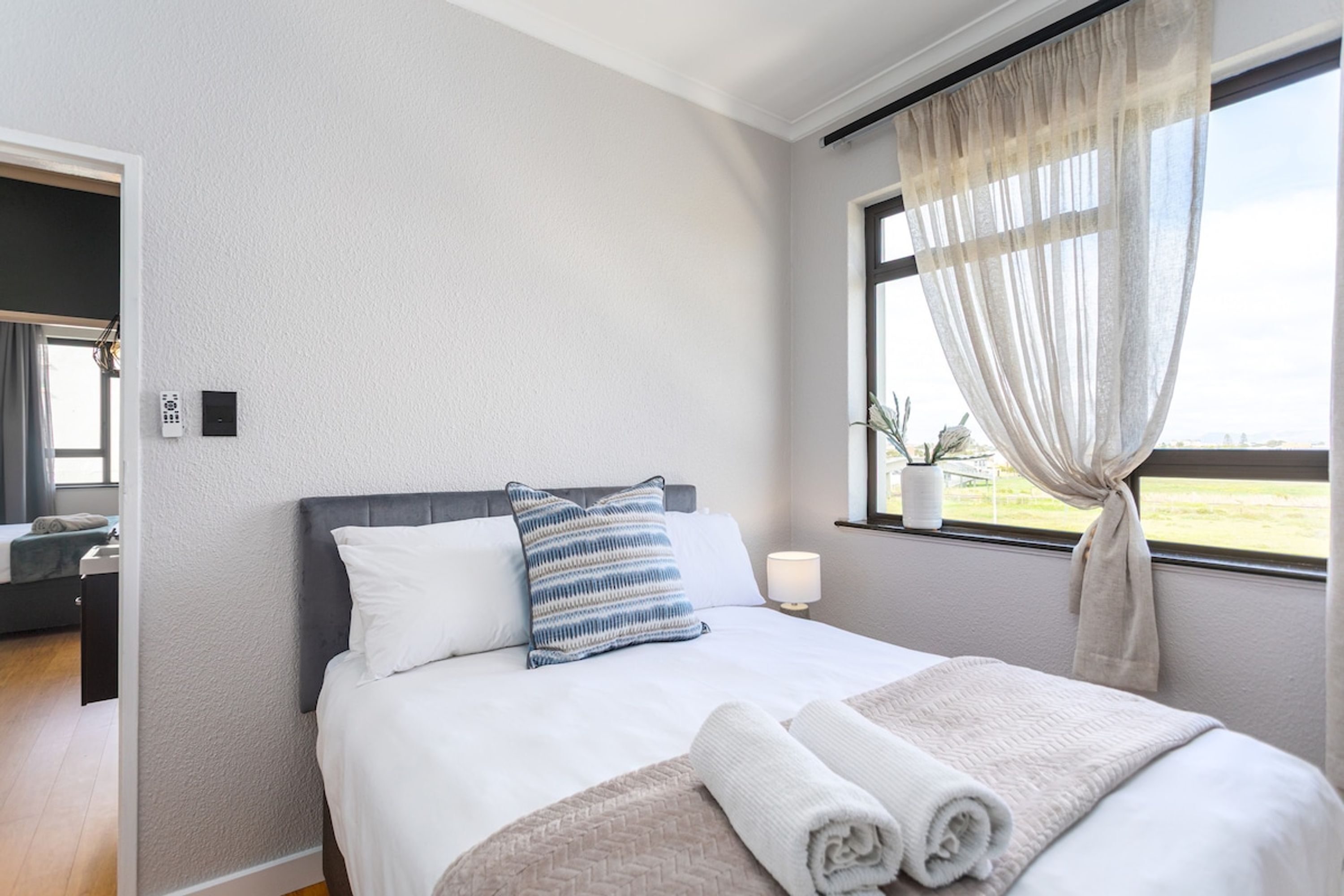 A hotel room with a bed, a window with curtains, a bedside table with a lamp, and a view of the outdoors.