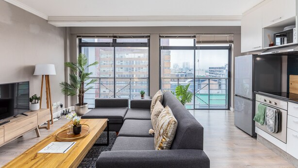 Ein modernes Wohnzimmer mit einem grauen Sofa, einem Holztisch und einem Fernseher. Ein Balkon bietet Blick auf Gebäude und die Stadtlandschaft.