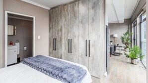 Ein Schlafzimmer mit großem Kleiderschrank, einem Bett mit strukturierter Decke, einem Nachttisch und Blick auf einen Flur mit Schreibtisch und Pflanzen.