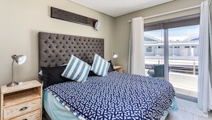 A bedroom with a large bed, a wooden nightstand, a bedside lamp, and a view of the outside balcony. - Seaside Village J9 by Ctha (Cape Town)