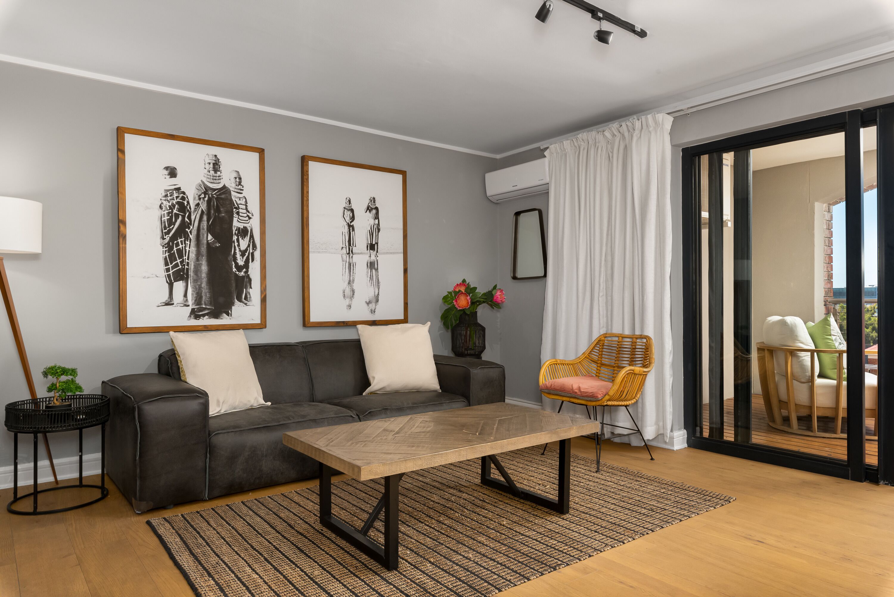 Un salon moderne avec un canapé gris, une table basse en bois et un fauteuil en osier. Deux photographies en noir et blanc, encadrées, sont accrochées au mur.