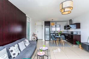 A modern living room with a grey sofa, two small round tables with plants, a dining area with a grey table and chairs, and a kitchen with dark cabinetry and stainless steel appliances. - Knightsbridge 702w by Ctha (Cape Town)