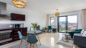 A modern apartment with a kitchen, dining area, and living room. It features a large window with a city view, a dining table with chairs, a sofa, and a small side table. - Knightsbridge 702w by Ctha (Cape Town)