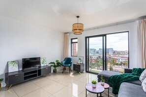 A modern living room with a flat-screen TV, a sofa, a chair, and a large window offering a city view. - Knightsbridge 702w by Ctha (Cape Town)
