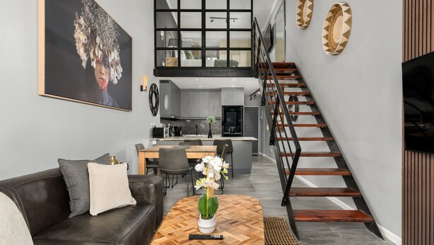 Ein modernes Wohnzimmer mit einem schwarzen Ledersofa, einem Holztisch und einem großen Gemälde an der Wand. Eine Treppe mit Holzboden führt auf eine obere Ebene.