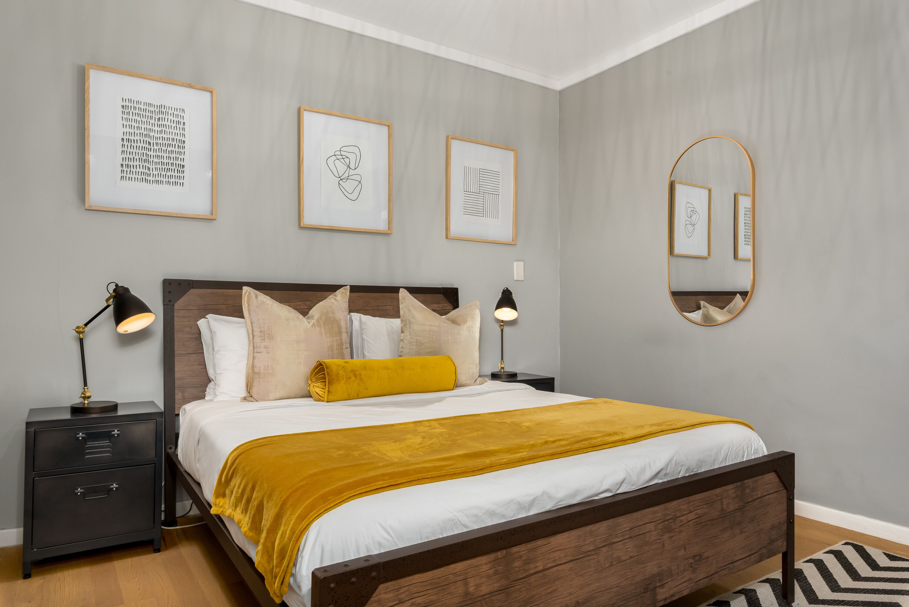 Un dormitorio con cama de madera, ropa de cama blanca, una manta color mostaza y una mesita de noche con lámpara. Hay obras de arte enmarcadas en la pared y un espejo sobre la cama.