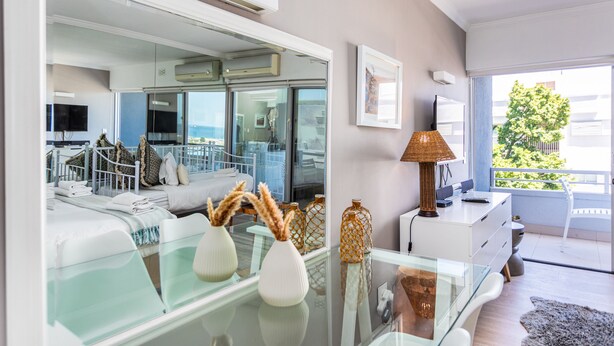 A modern hotel room with a glass table, white furniture, a large mirror, and a view of the outdoors. - First Beach 203 by Ctha (Cape Town)