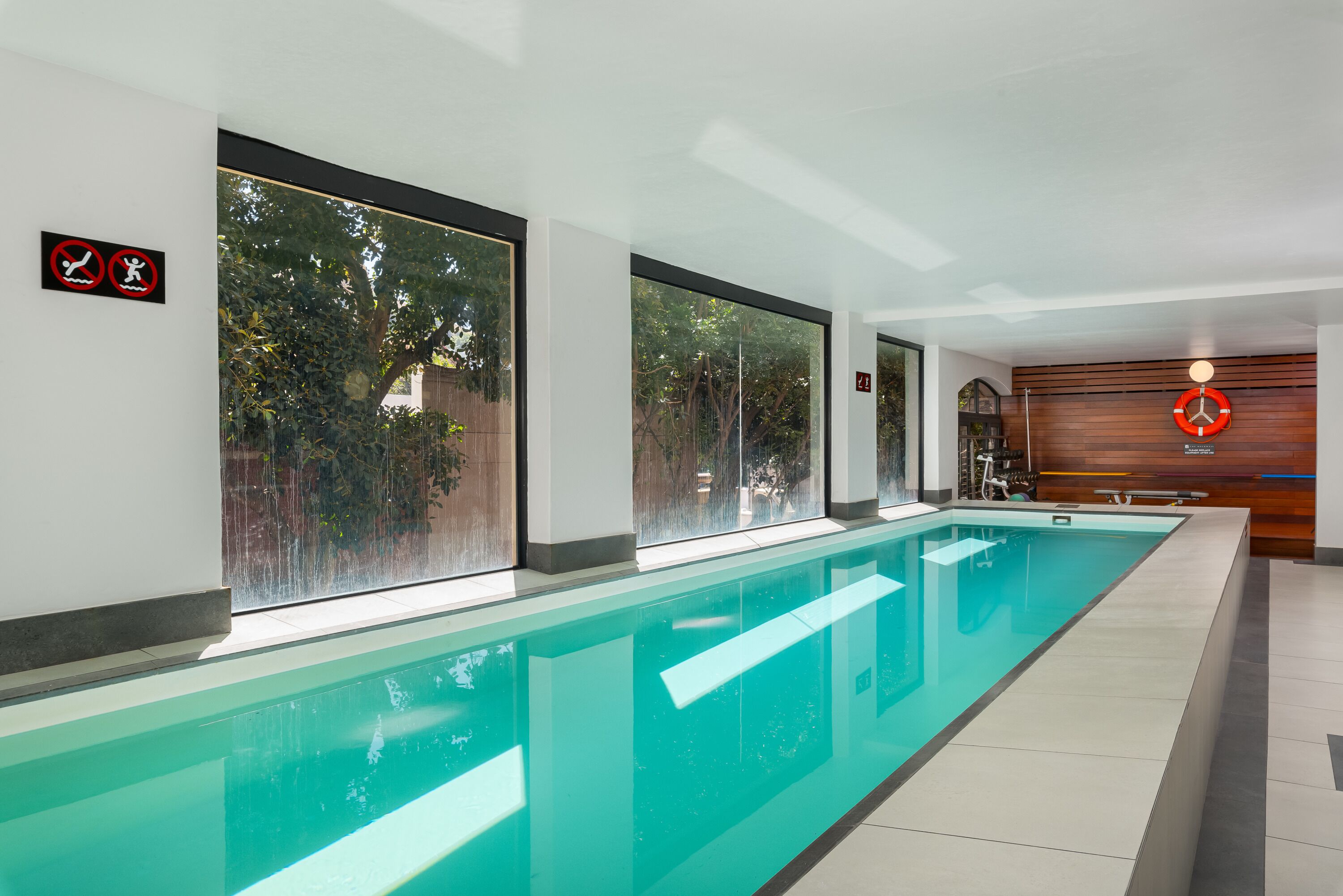 A long indoor swimming pool with clear glass walls, large windows, and a wooden wall with a clock.