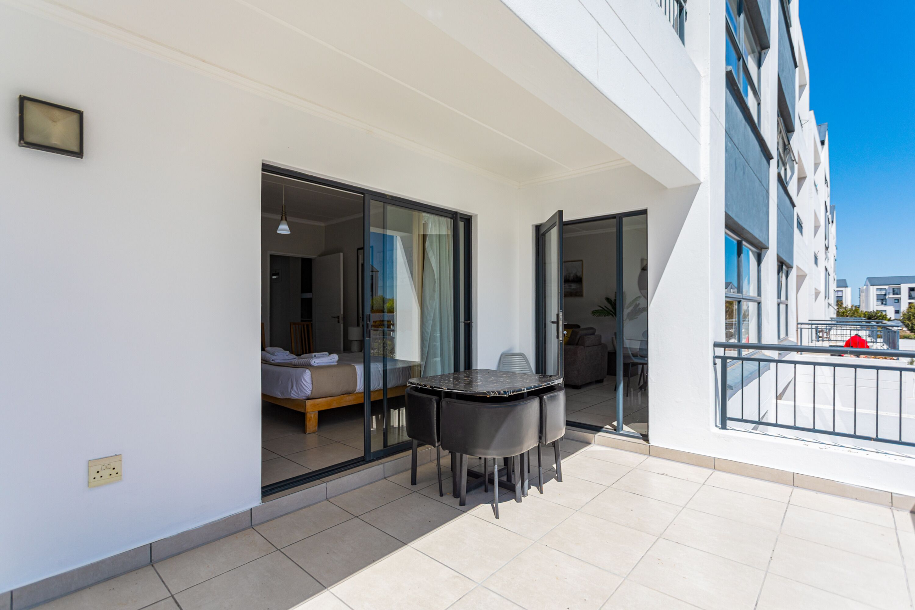 A balcony with a table and chairs, a bed, and a glass door leading to a room.