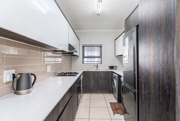 A modern kitchen with white cabinets, a stainless steel kettle, a gas stove, a built-in oven, and a refrigerator. - The Sandown 178 by Ctha (Cape Town)
