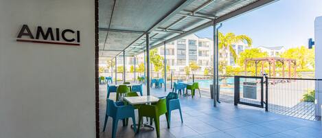 An outdoor patio area with tables and chairs, a sign reading "AMICI", and a view of buildings and trees in the background.