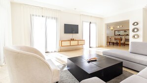 Un salon moderne avec un canapé, un fauteuil et une table basse. Un téléviseur à écran plat est fixé au mur, et on aperçoit un coin repas à l’arrière-plan.