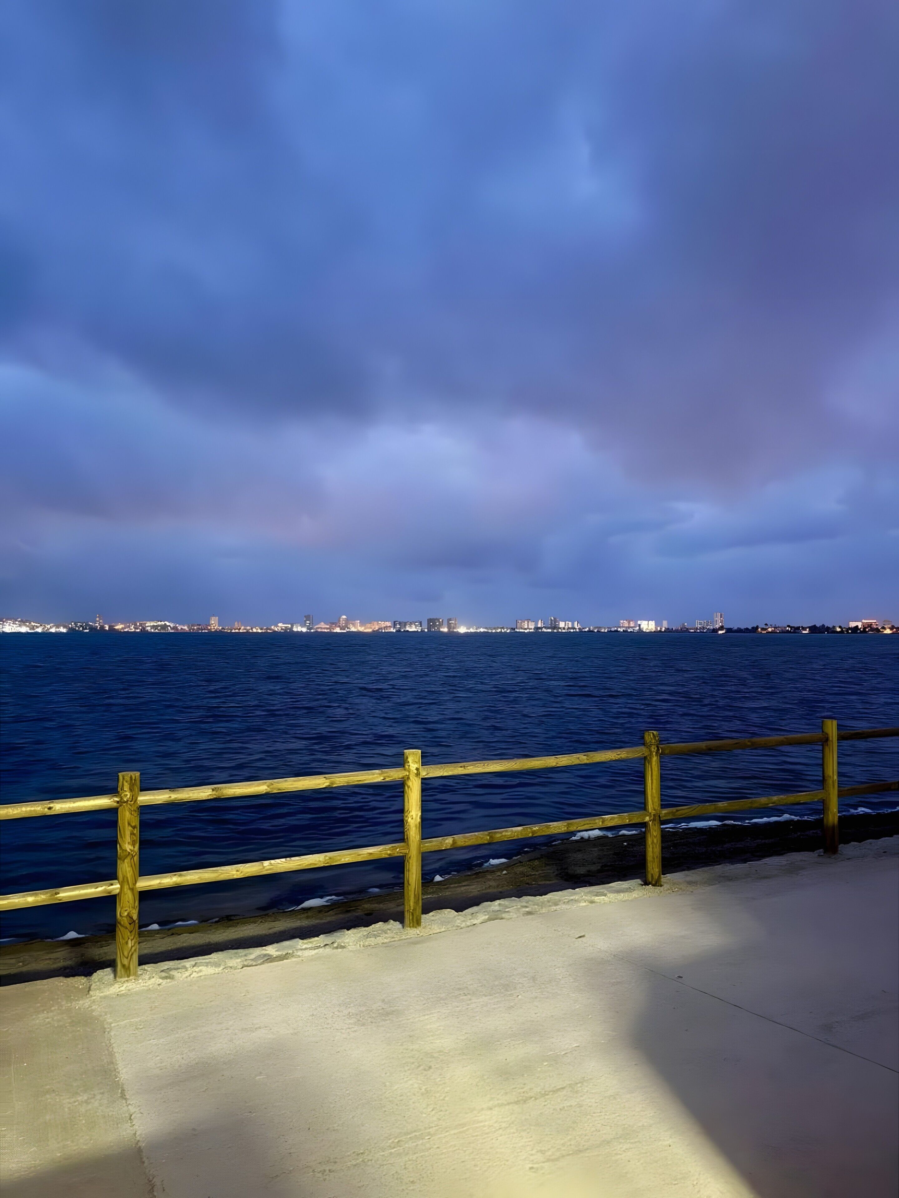 A waterfront view with a wooden railing, a concrete walkway, and a distant city skyline.