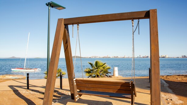 A wooden swing set by a sandy beach with a clear view of the sea and distant city skyline.