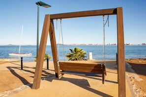 A wooden swing set by a sandy beach with a clear view of the sea and distant city skyline.