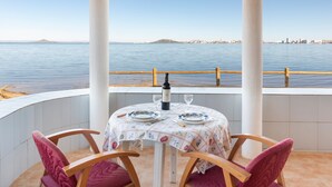 A table with a bottle and glasses, two chairs, a view of the sea, and a cityscape in the background.