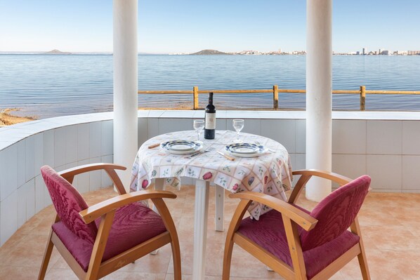 A table with a bottle and glasses, two chairs, a view of the sea, and a cityscape in the background.