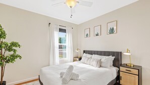 Una cama bien hecha con sábanas blancas, una mesita de noche con lámpara y un ventilador de techo.